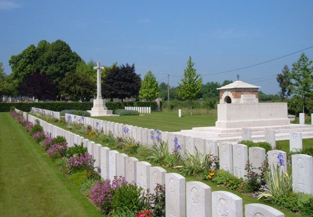 Photo d’un cimetière militaire contenant deux rangées de pierres tombales entourées de fleurs. Une grande croix et le derrière d’un monument sont visibles en arrière-plan.