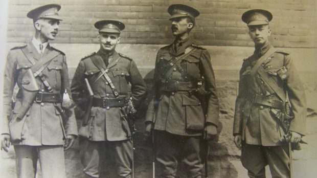 Quatre officiers militaires hommes debout en train de parler devant un bâtiment. Le deuxième en partant de la droite tient les mains derrière son dos. Ils portent tous des étuis pour pistolet et des épées.