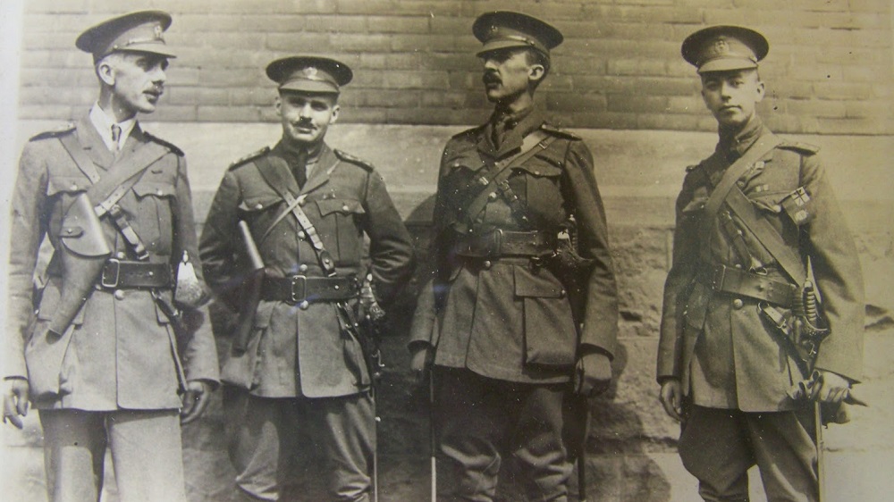 Quatre officiers militaires hommes debout en train de parler devant un bâtiment. Le deuxième en partant de la droite tient les mains derrière son dos. Ils portent tous des étuis pour pistolet et des épées.