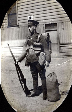 L’image d’un soldat debout dans un passepartout ovale. Il tient son fusil dans la main droite. Un sac marin se trouve à côté de son pied gauche. On peut voir un bâtiment à l’arrière-plan.