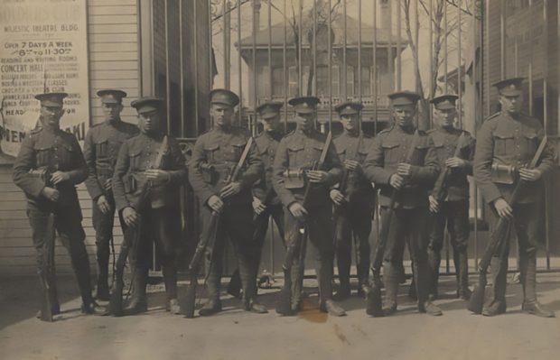 Dix soldats, tous portant une casquette et tenant chacun un fusil. Ils sont debout devant un portail; on entrevoit sur la droite l’enseigne d’un théâtre.