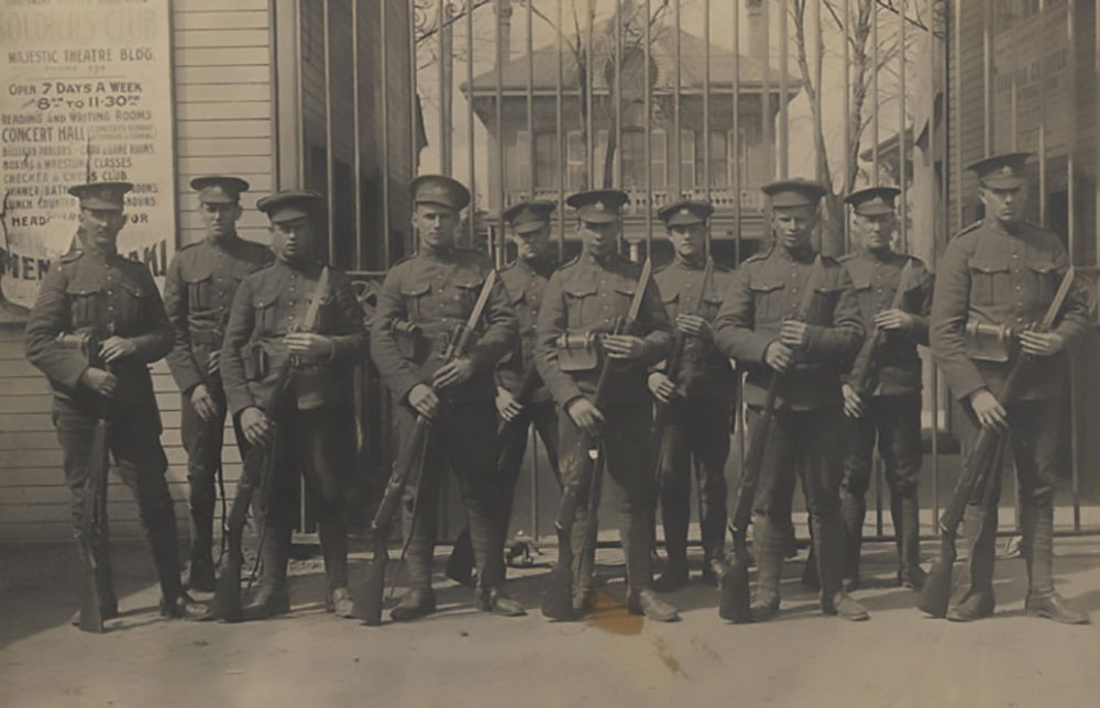 Dix soldats, tous portant une casquette et tenant chacun un fusil. Ils sont debout devant un portail; on entrevoit sur la droite l’enseigne d’un théâtre.