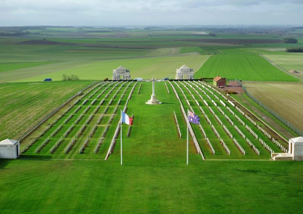 Photo d’un cimetière militaire à la campagne, contenant plusieurs rangées de pierres tombales. En avant-plan, on peut remarquer les drapeaux français et australien.