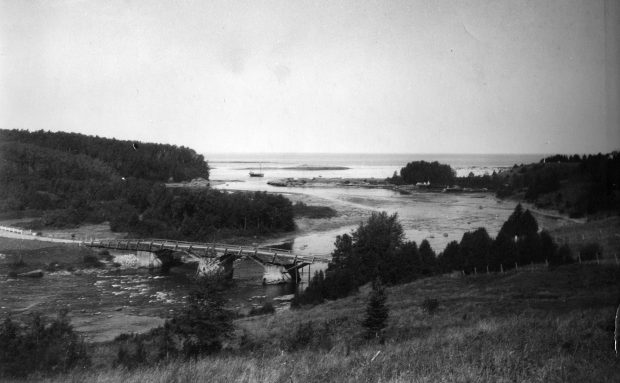 Photographie du vieux pont de bois de Grand-Métis vers 1925. Le pont est situé à environ 1 km de l’embouchure de la rivière Metis. Les deux côtés de la rivière présentent un paysage forestier et les aménagements du quai. Une goélette est amarrée à l’embouchure de la rivière et laisse entrevoir l’étendue du fleuve Saint-Laurent. 