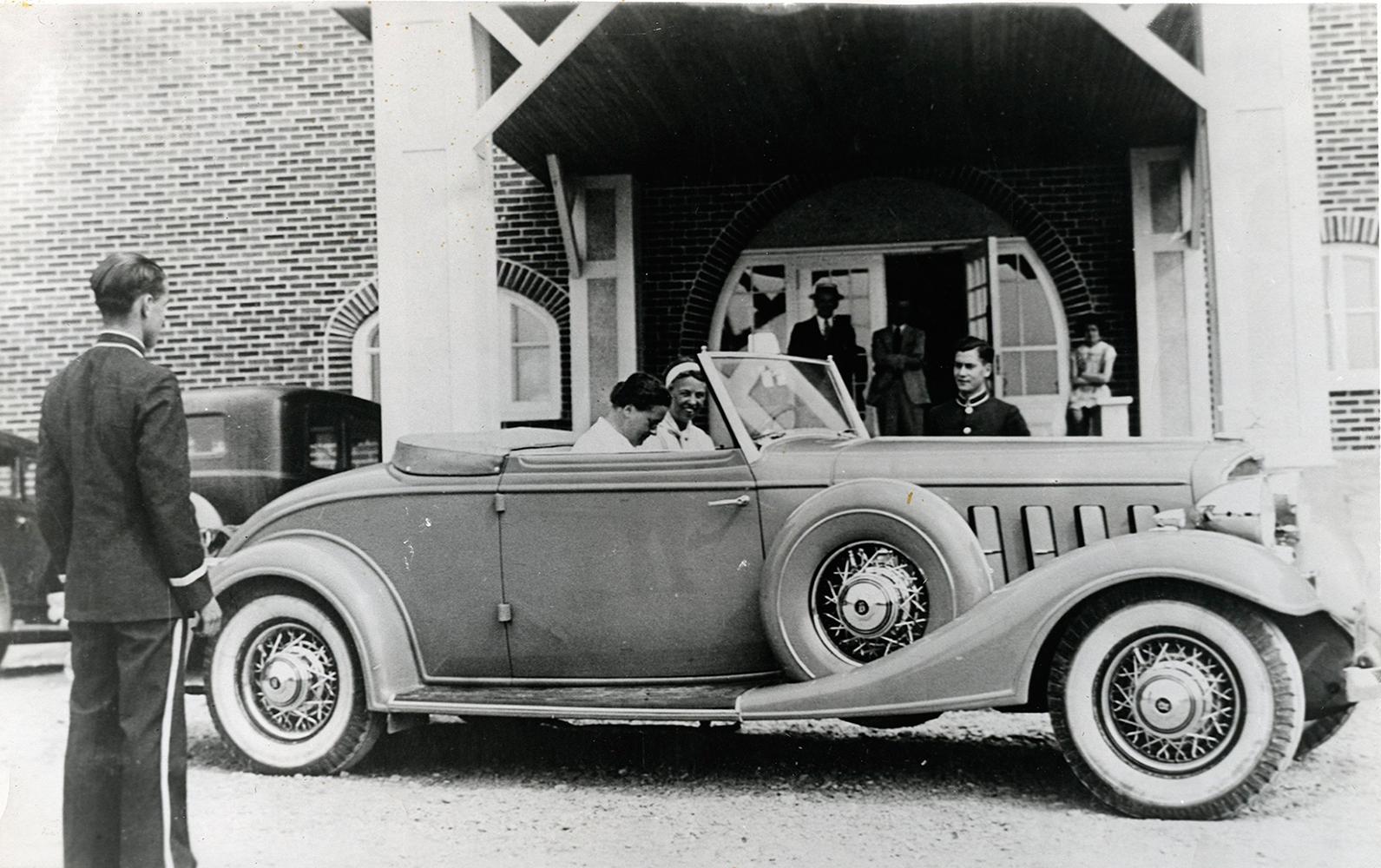 Photographie noir et blanc. Dans une Buick convertible, deux femmes, Eleanor Roosevelt et son amie Lorena Hickok s’apprêtent à partir. La voiture est stationnée à l’avant de l’hôtel La Belle Plage de Matane. Un valet de l’hôtel attend leur départ. 