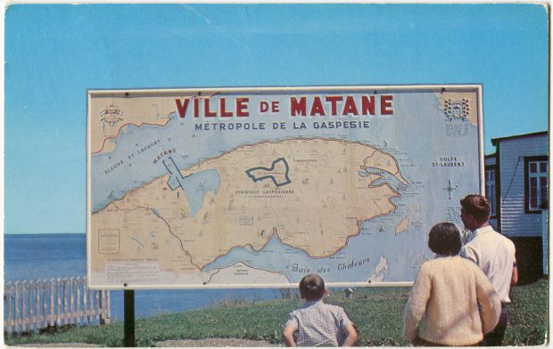 Un homme, une femme et un enfant observent une carte de la Gaspésie. La carte et une affiche d’une dimension approximative de 4 pieds de haut par 8 pieds de large. Installée à l’extérieur à proximité du phare de Matane.  On peut y lire : Ville de Matane Métropole de la Gaspésie.