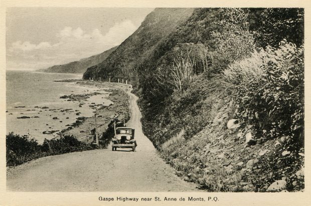 Carte postale présentant une voiture sur le boulevard Perron vers 1930, alors qu’il était en gravier. Le boulevard Perron longe le pied d’une montagne abrupte et la berge du fleuve Saint-Laurent près de Sainte-Anne-des-Monts.