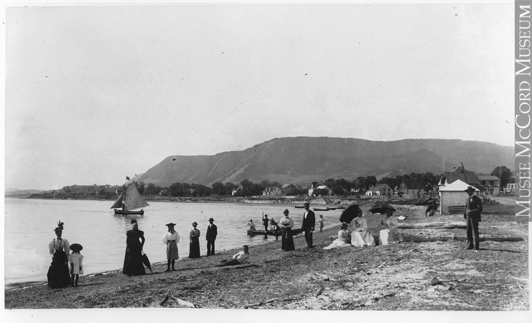 Photographie en noir et blanc d’une plage. Une douzaine de personnes posent, les hommes en habits et les femmes en robes longues, certaines portant des ombrelles. Un voilier et d’autres petits bateaux quittent la rive. En arrière-plan quelques maisons rustiques au pied d’une montagne.