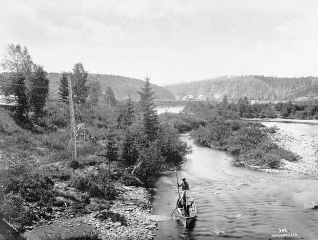 Paysage forestier traversé par une rivière. Au bas de la photographie deux hommes autochtones manient un canot à l’aide de pôles. Sur la berge de la rivière on aperçoit un tipi et au loin quelques maisons rustiques au pied deux collines.