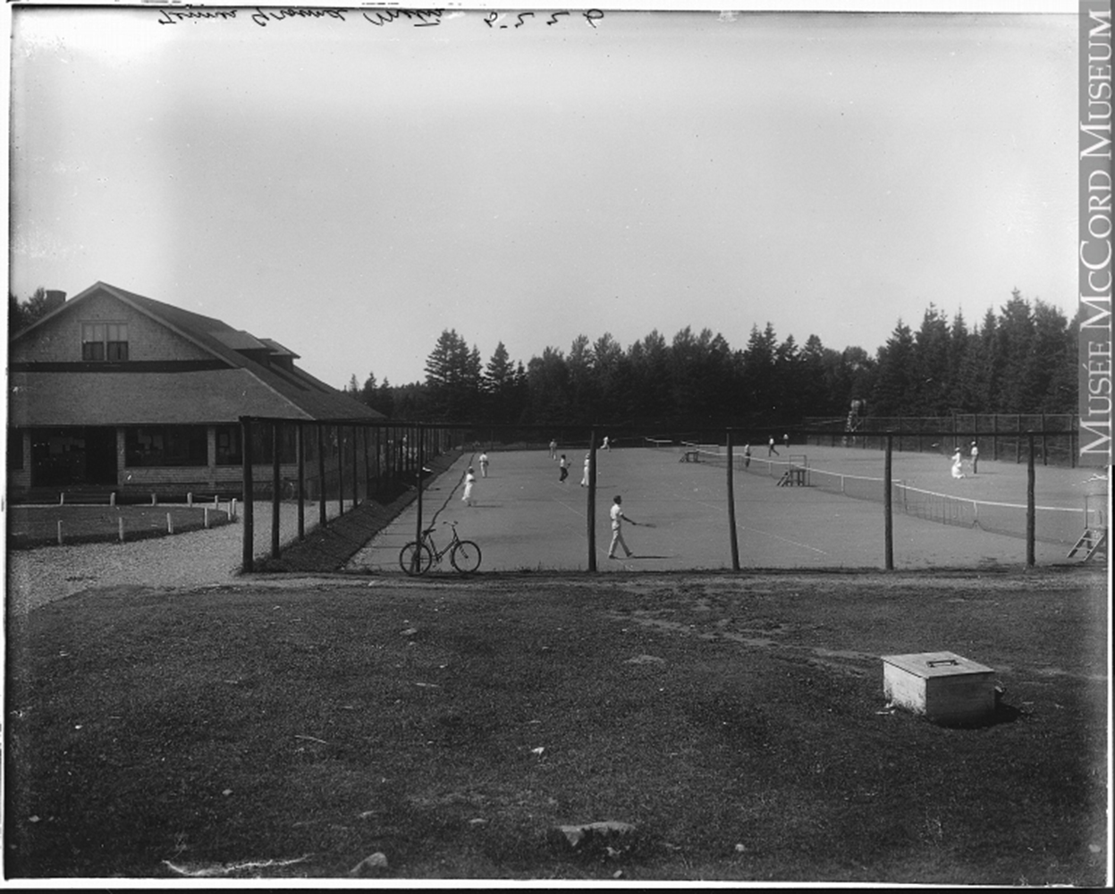 Photographie noir et blanc du terrain de tennis de Métis-sur-Mer. Une haute clôture entoure le terrain de tennis.  Des hommes, vêtu d’un pantalon et d’un chandail blanc, et des femmes, vêtu d’une longue jupe blanche d’une chemise blanche à manche longue et coiffée d’un chapeau, jouent au tennis. Du côté gauche, on aperçoit le bâtiment du club de golf Cascade.