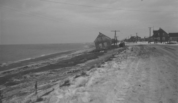 Photographie noir et blanc d’une maison à deux étages située à l’entrée longeant le fleuve. La maison se trouve sur la plage, étrangement inclinée d’un côté, il semble qu’elle ait été déplacée de ce côté de la route par les vagues. 