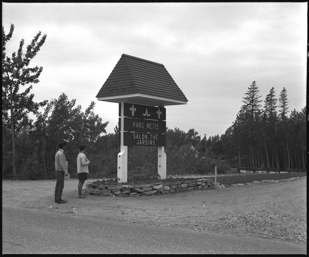 Photographie noir et blanc de l’entrée du Parc Métis. Un homme et une femme observent l’affiche située à l’entrée du site, près de la route 132. 