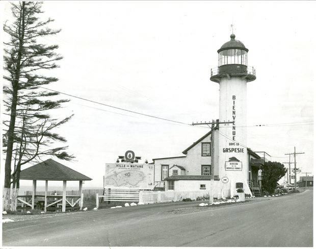 Photographie noir et blanc du phare de Matane adapté en bureau d’information touristique. Le boulevard Perron occupe le premier plan de la photographie. Le phare et la maison du gardien sont situés du côté droit de l’image. Sur le phare il est écrit : Bienvenue dans la Gaspésie. Une affiche sur le phare mentionne : Berceau de la Nouvelle-France. Une immense carte de la Gaspésie est affichée près du phare. 