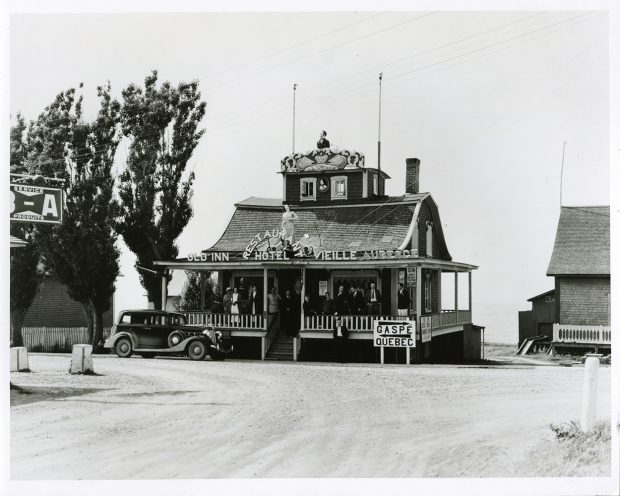 Une douzaine de personnes posent sur la galerie d’une maison à deux étages en bardeaux de cèdres. La maison est décorée de deux figures de proue. L’une, le buste d’un homme, a été installée sur le dessus d’une tour d’observation qui dépasse du centre de la toiture. La deuxième, une sculpture grandeur nature d’une femme, vêtue d’une robe blanche légère et installée sur le toit de la galerie au-dessus de la porte principale. Des lettres ont été fixées sur cette même toiture pour indique le nom de l’auberge. On peut y lire : Old Inn Hotel Restaurant Hotel la Vieille Auberge. 