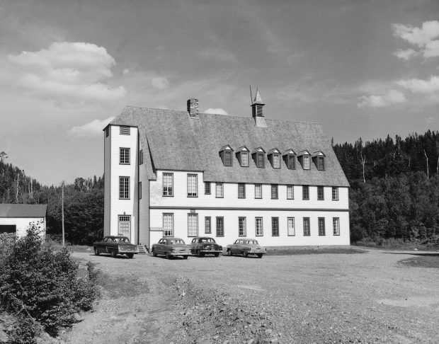 Photographie noir et blanc du Gîte du Mont-Albert en 1950.  Le gîte est un imposant bâtiment rectangulaire de 3 étages au cœur d’une forêt de conifères. L’angle prononcé du toit à double pente lui donne un style architectural qui oscille entre celui d’un chalet suisse et d’un manoir. 