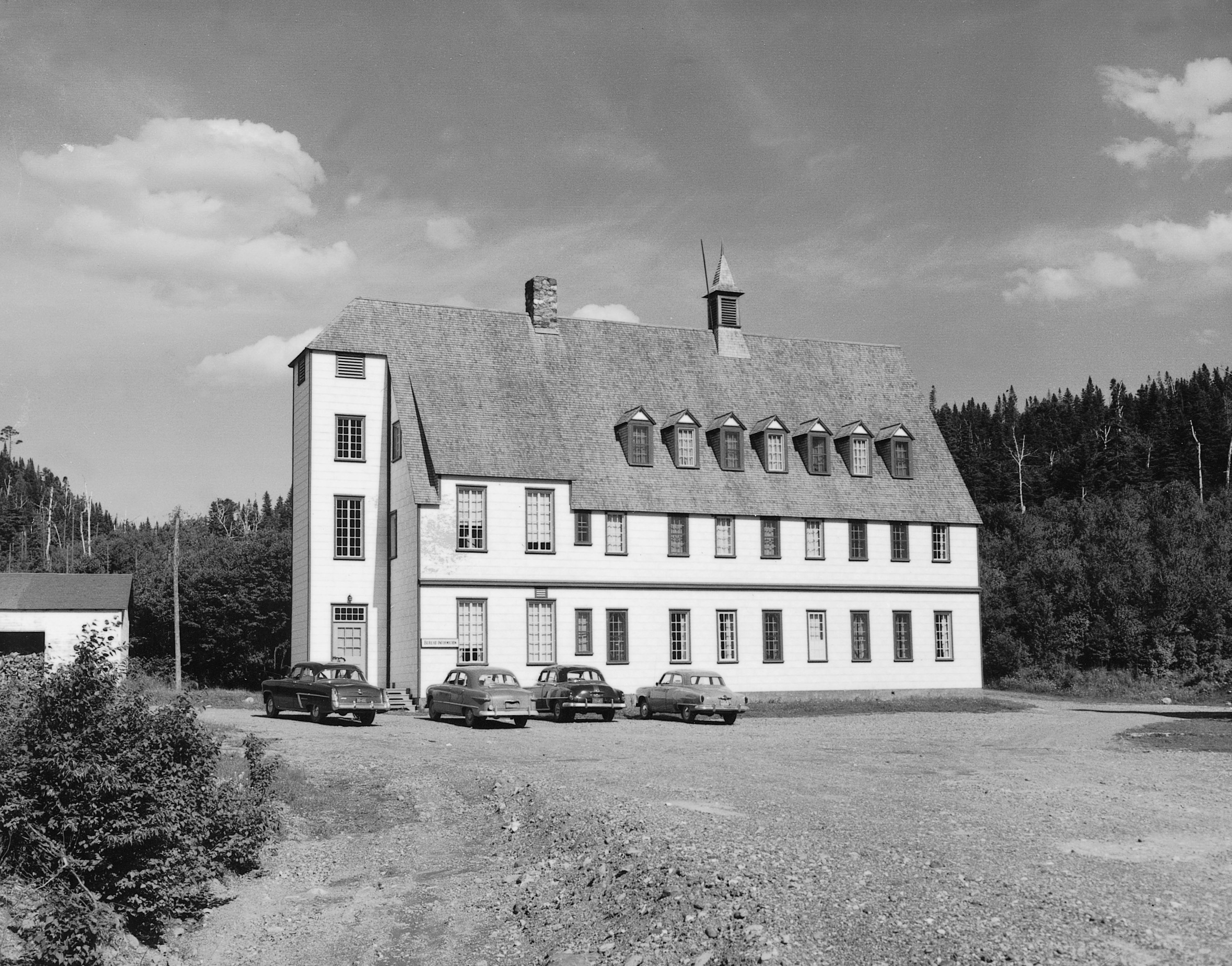 Photographie noir et blanc du Gîte du Mont-Albert en 1950.  Le gîte est un imposant bâtiment rectangulaire de 3 étages au cœur d’une forêt de conifères. L’angle prononcé du toit à double pente lui donne un style architectural qui oscille entre celui d’un chalet suisse et d’un manoir. 