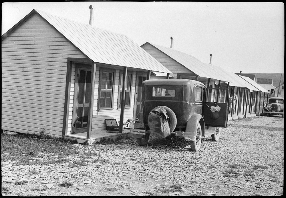 Photographie noir et blanc d’une série de cabines touristiques à Percé. Une voiture des années 1933 est stationnée devant la première cabine. 