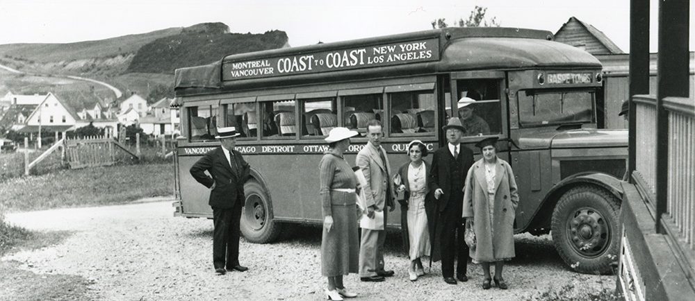 Photographie noir et blanc d’un groupe de personnes devant un autobus voyageur de 1932. Derrière l’autobus, on peut apercevoir quelques maisons du village de Percé et les montagnes qui forment la côte. Sur l’autobus il est écrit : Montreal Vancouver Coast to Coast New York Los Angeles.