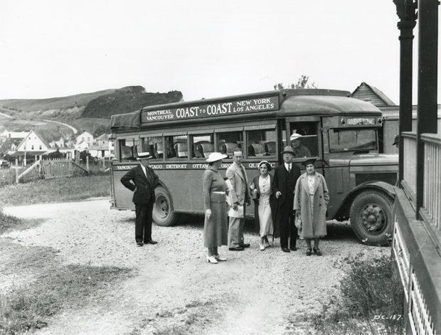 Photographie noir et blanc d’un groupe de personnes devant un autobus voyageur de 1932. Derrière l’autobus, on peut apercevoir quelques maisons du village de Percé et les montagnes qui forment la côte. Sur l’autobus il est écrit : Montreal Vancouver Coast to Coast New York Los Angeles.
