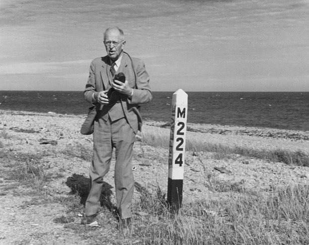Portrait de George Parmenter. L’homme âgé, habillé en veston cravate, se tient debout, son appareil photo à la main, près d’un poteau blanc sur lequel il est inscrit M224. En arrière-plan la plage de Sainte-Flavie et la mer.
