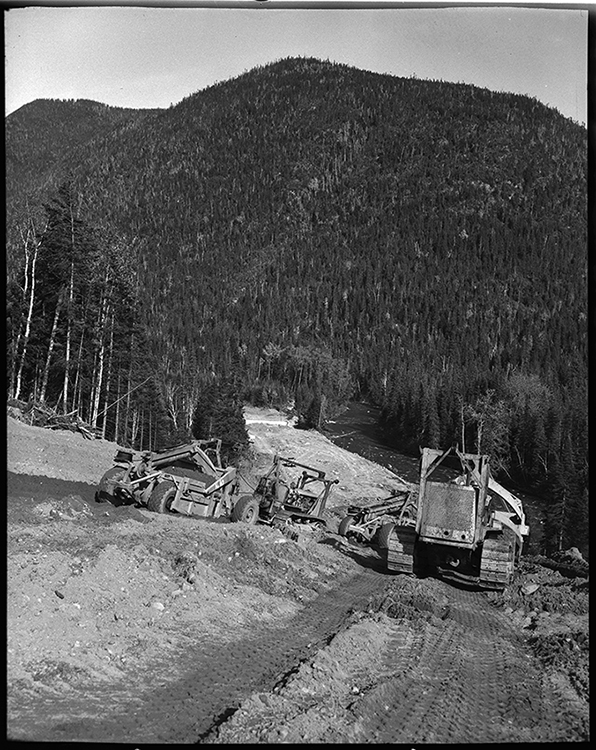 Photographie noir et blanc des travaux sur le chemin Trans-Gaspésien. Dans un paysage forestier, sur une pente abrupte, des travailleurs de la construction manipulent de la machinerie afin de faire les travaux de préparation d’aménagement de la route. 