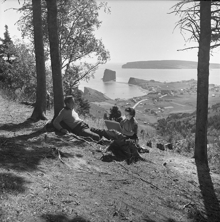 Photographie noir et blanc de l’artiste Suzanne Guité à l’œuvre d’un dessin avec comme modèle son mari, l’artiste Alberto Tommi. Le couple est assis au pied d’un arbre au sommet d’une montagne. Derrière eux, un magnifique paysage se dépolit, on aperçoit au bas de la montagne le village de Percé ainsi que le rocher Percé. 