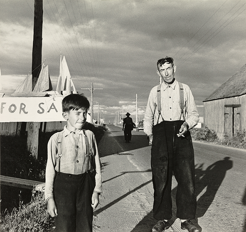 Photographie noir et blanc d’un homme et d’un jeune garçon sur le bord de la route. L’homme et l’enfant sont vêtus d’une chemise blanche et d’un pantalon noir retenu par des bretelles. Derrière le garçon des bateaux de bois sont déposés sur le dessus d’une affiche sur laquelle il est inscrit « For Sale » à vendre. 