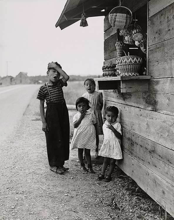 Portrait noir et blanc de quatre enfants Mi`gmaq au-devant d’un kiosque routier de ventre de panier en vannerie, sur le bord de la route.