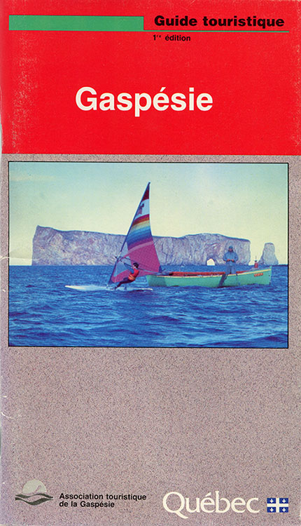 Couverture de la première édition du guide touristique de la Gaspésie. Au centre de la page se trouve une photographie d’un pêcheur se tenant debout dans une chaloupe et d’un homme pratiquant le sport de la planche à voile. En arrière-plans, nous apercevons le Rocher Percé.