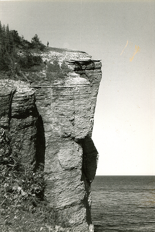 Photographie noir et blanc d’une falaise rocheuse d’une hauteur impressionnante. Au sommet de celle-ci on distingue la minuscule silhouette d’un homme. En arrière-plan le golfe du Saint-Laurent.  