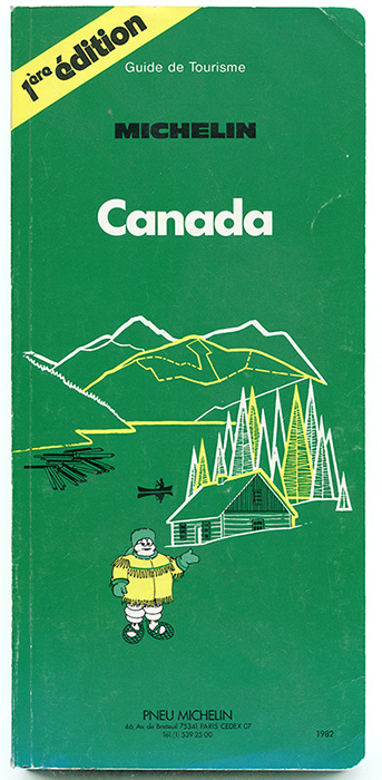 Page couverture de la première édition du guide Michelin. Sur un fond vert foret, la mascotte des pneus Michelin est vêtue d’un manteau à frange. Il porte des raquettes un chapeau en fourrure. Derrière lui un chalet en bois rond dans une forêt de conifères près d’un lac sur lequel il se trouve un canot et des billots de bois. En arrière-plan, les montagnes se succèdent.