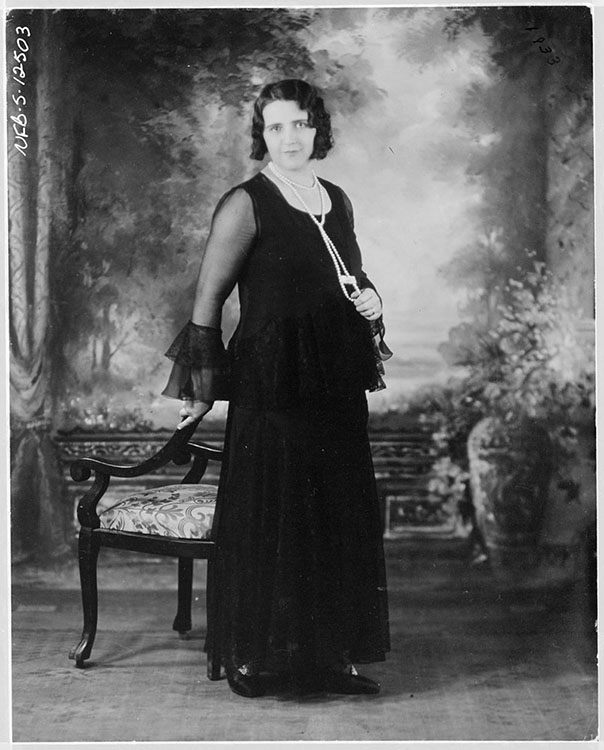 Portrait studio noir et blanc de la chanteuse la Bolduc. Elle porte d’une robe longue noire est debout, près d’une chaise style victorienne, au centre de la photographie devant un fond peint du style classique.  