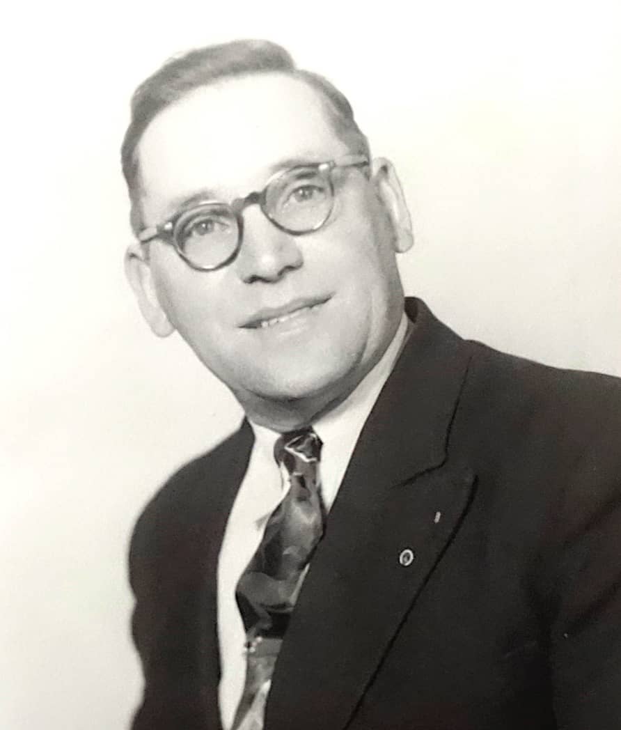 John Dominie portant des lunettes et vêtu d’un costume et d’une cravate