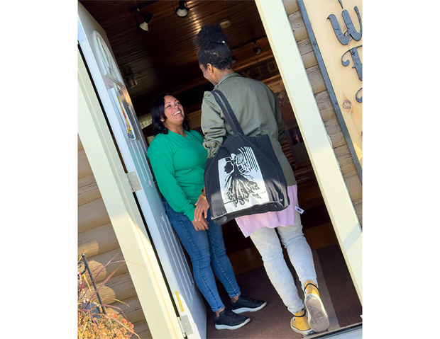 Émilie Jabouin et Michelle Robbins se saluent dans l’embrasure d’une porte. Michelle, vêtue de vert, se tient à l’intérieur en souriant; Émilie, portant un sac fourre-tout noir avec un motif blanc, est debout à l’entrée.