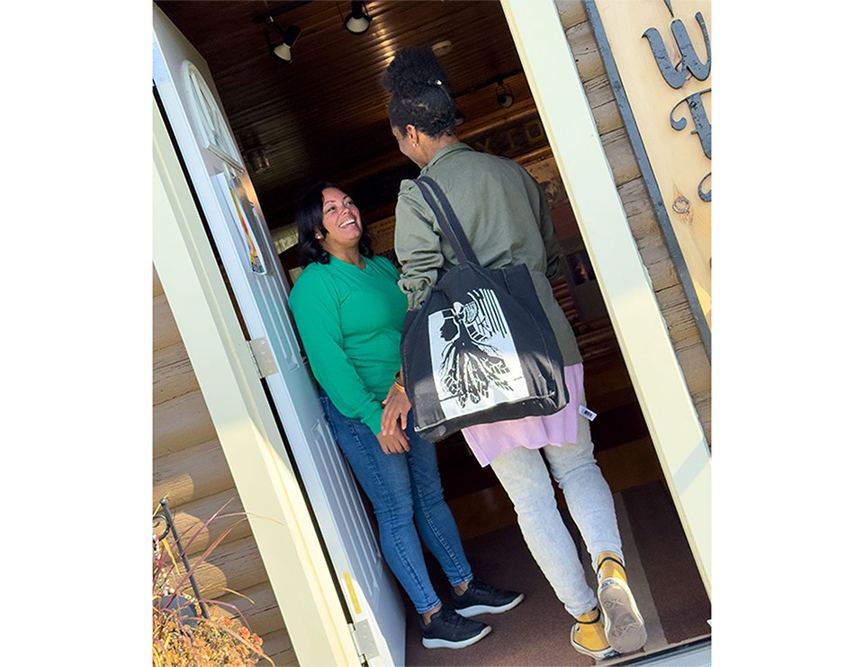 Émilie Jabouin et Michelle Robbins se saluent dans l’embrasure d’une porte. Michelle, vêtue de vert, se tient à l’intérieur en souriant; Émilie, portant un sac fourre-tout noir avec un motif blanc, est debout à l’entrée.
