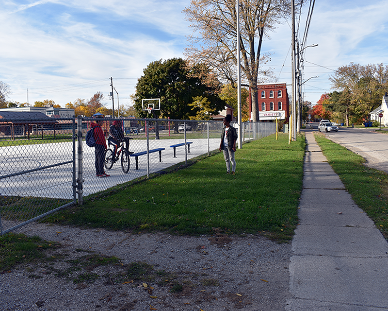 Une scène de quartier montrant deux jeunes hommes avec un vélo sur un terrain de basket, tandis qu'Emily marche sur le sentier extérieur à proximité d'une épicerie locale.