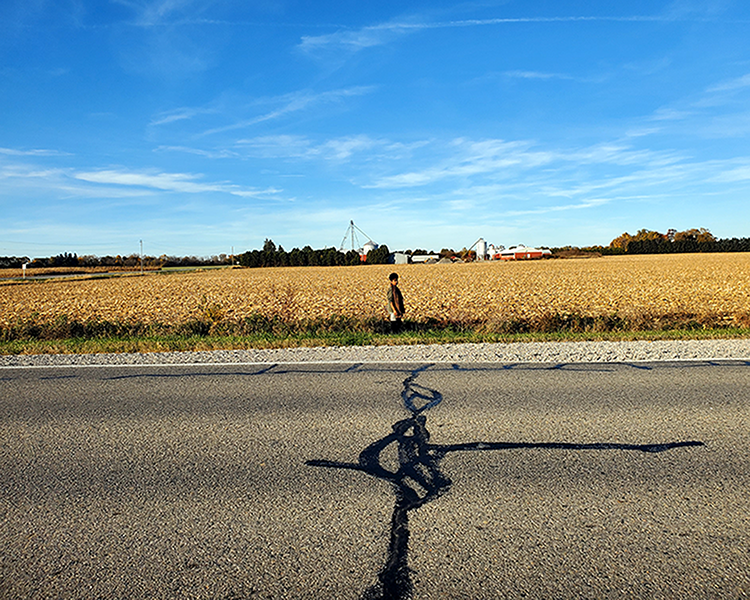 Emily se tient près d’un champ récolté le long d’une route rurale, avec des bâtiments de ferme et des silos visibles à l’arrière-plan sous un ciel bleu.