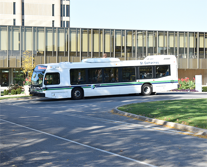 Un autobus urbain blanc portant l'inscription « St. Catharines » sur le côté circule sur une route courbée devant un bâtiment aux grandes fenêtres vitrées à l’Université Brock.