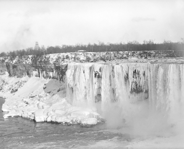 Une photo en noir et blanc des chutes Niagara en hiver, avec de grandes sections de la cascade et du paysage environnant recouvertes de glace et de neige. Une brume s’élève des chutes, et des arbres bordent l’arrière-plan.