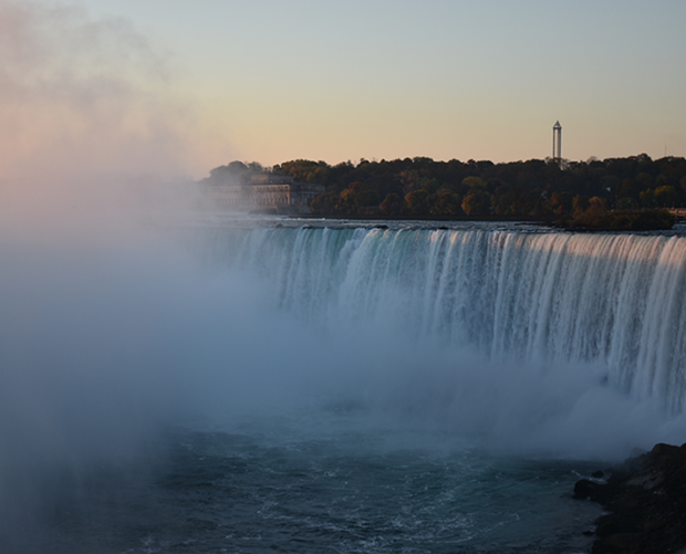  Une vue large des chutes Niagara où l’eau se précipite par-dessus le rebord, la brume s’élève, et l’horizon bordé d’arbres s’étend sous un ciel dégagé au coucher du soleil.