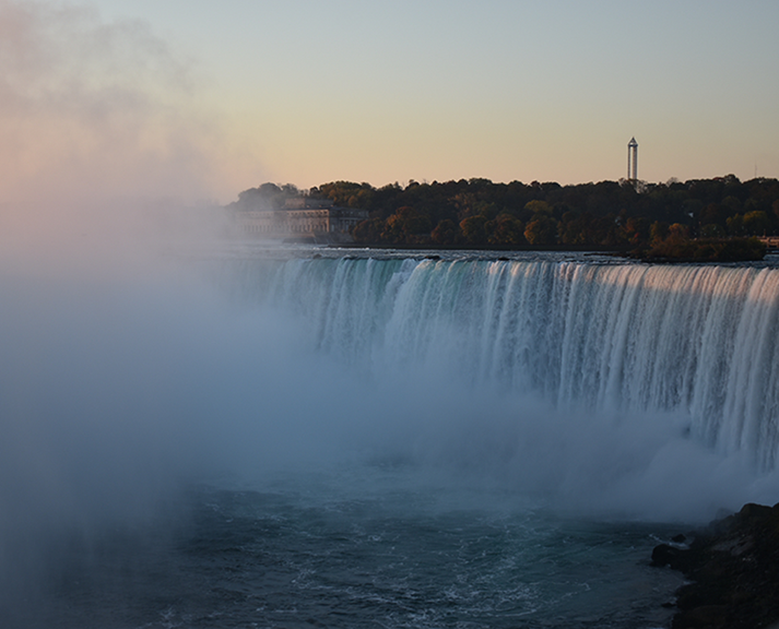  Une vue large des chutes Niagara où l’eau se précipite par-dessus le rebord, la brume s’élève, et l’horizon bordé d’arbres s’étend sous un ciel dégagé au coucher du soleil.