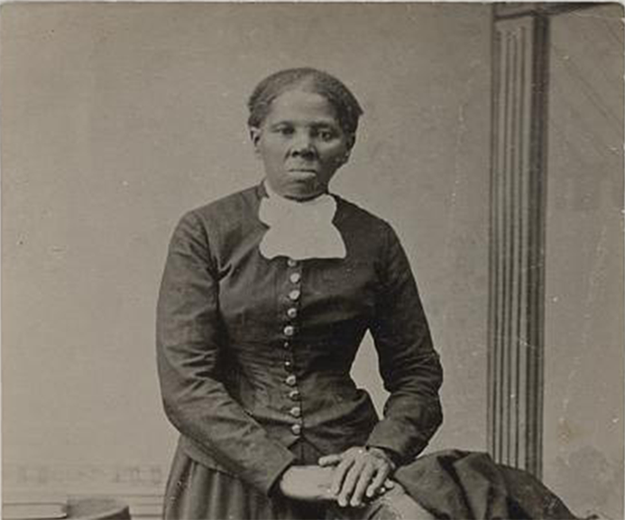 Harriet Tubman porte une longue robe foncée boutonnée. Elle se tient debout à côté d’une chaise recouverte d’un manteau, une main posée sur le dossier.