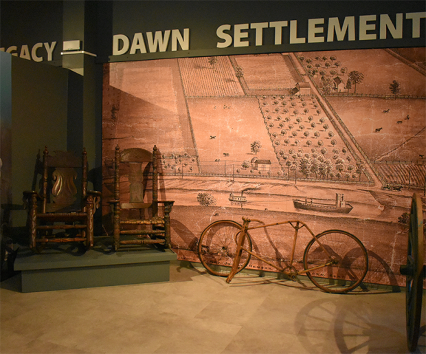 Au Musée Josiah Henson de l’histoire afro-canadienne, une exposition met en valeur deux fauteuils berçants en bois, une vieille bicyclette et une murale illustrant une colonie rurale avec des champs et des bâtiments. Les mots Dawn Settlement apparaissent au-dessus de la murale.