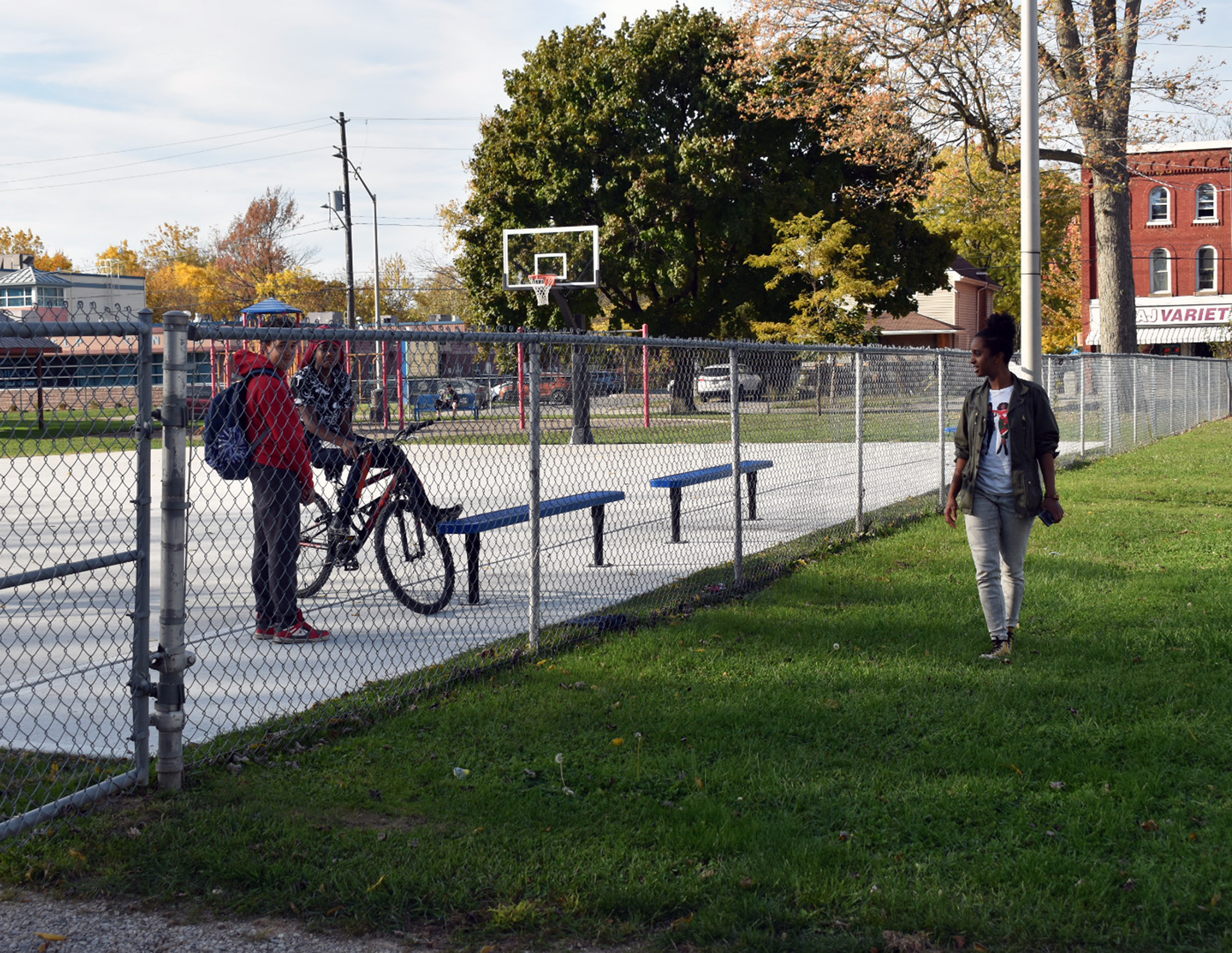 Une scène de quartier montrant deux jeunes hommes avec un vélo sur un terrain de basket, tandis qu'Emily marche sur le sentier extérieur à proximité d'une épicerie locale.