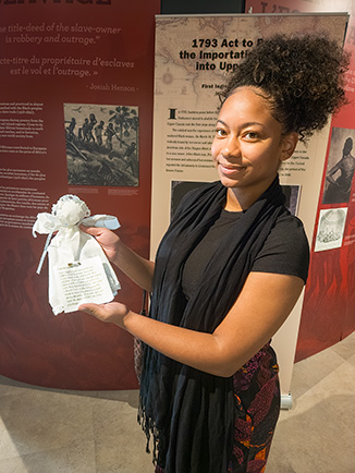 À l’intérieur de l’exposition du Musée Josiah Henson de l’histoire des Afro-Canadiens, une jeune femme sourit en tenant une poupée artisanale fabriquée avec des mouchoirs, accompagnée d’une étiquette. Derrière elle, des panneaux présentent la Loi de 1793 visant à limiter l’esclavage dans le Haut-Canada ainsi que l’histoire de l’esclavage.