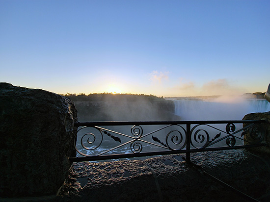 Vue sur les chutes du Niagara au lever du soleil, avec la brume qui s’élève au-dessus de la cascade et la lumière matinale illuminant une rambarde en fer forgé.