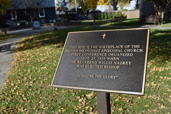 Une plaque en bronze montée sur un poteau dans un parc à Chatham-Kent, soulignant le lieu de naissance de l’Église épiscopale méthodiste britannique. Des feuilles jonchent l’herbe autour, et un bâtiment historique est visible à l’arrière-plan. Photo de Blessing Ogunyemi.