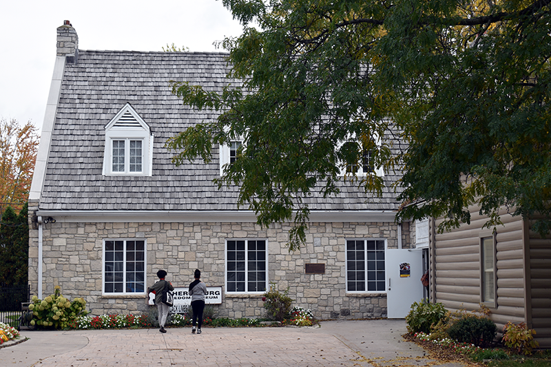 Deux chercheurs se tiennent devant l’Amherstburg Freedom Museum, lisant un panneau d’information près de l’entrée du bâtiment historique en pierre. Le musée est entouré de fleurs et de grands arbres aux couleurs automnales.