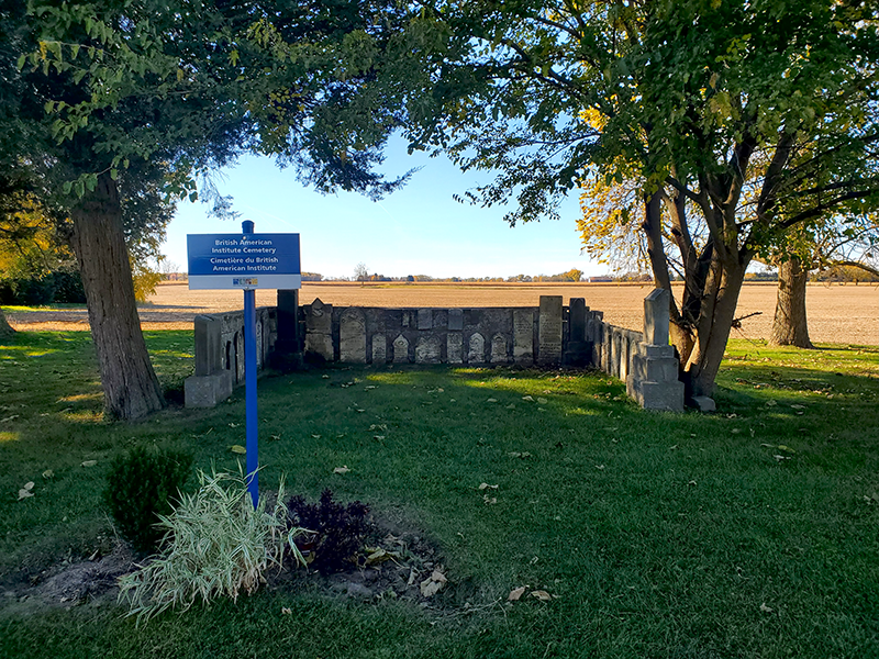 Vue du cimetière de l’Institut britannique américain au Musée Josiah Henson de l’histoire afro-canadienne. Un panneau historique bleu se dresse sur une zone herbeuse entre deux grands arbres, avec un petit enclos de vieilles pierres tombales derrière lui et des champs ouverts à l’horizon.