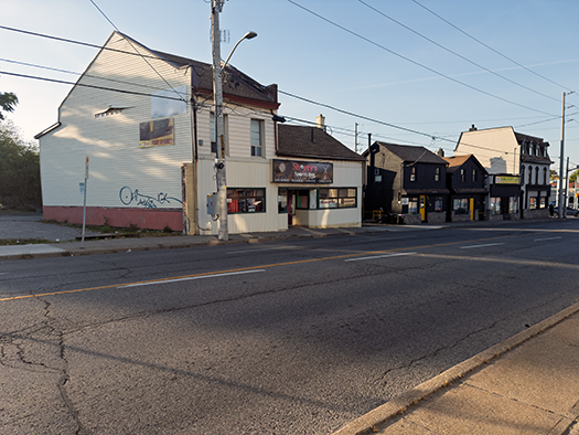 Vue de la rue Geneva à St. Catharines, montrant une rangée de petits commerces et maisons bordant la route sous une lumière de début ou de fin de journée. Des lignes électriques traversent la scène, et la rue semble calme. Photo de Blessing Ogunyemi.